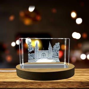 Op de afbeelding: Een helder kristallen blok met een 3D-geëtst kasteelontwerp, verlicht door een houten basis. Het kasteel heeft twee torens, een centrale poort en bomen. De basis is donkerbruin.