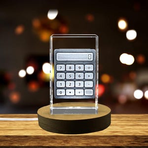 Op de afbeelding: Een heldere glazen kubus met een gegraveerde zilveren rekenmachine erin. De kubus staat op een donkerbruine houten basis.