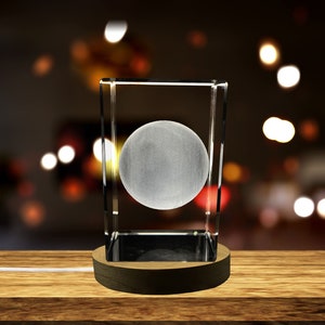 Könnte beinhalten: Ein klarer Glaswürfel mit einer mattierten Kugel im Inneren, der auf einem Holzsockel steht. Der Würfel wird von innen beleuchtet und erzeugt so ein sanftes Leuchten.