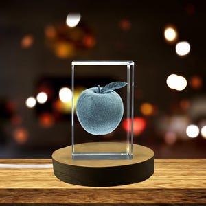 May include: A clear glass cube with a 3D etched image of a blue apple on a wooden base. The base has a cord attached to it.