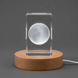 Könnte beinhalten: Ein klarer, rechteckiger Glasblock mit einer lasergravierten weißen Kugel in der Mitte, montiert auf einem runden, hellen Holzsockel. Das Objekt verfügt über ein weißes Stromkabel, das seitlich aus dem runden Holzständer herausragt.
