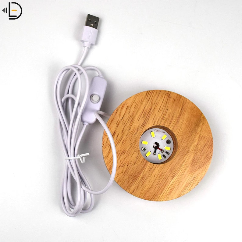K&ouml;nnte beinhalten: Ein Holzsockel mit einem runden Ausschnitt f&uuml;r ein Licht. Das Licht hat ein wei&szlig;es USB-Kabel mit einem Inline-Schalter. Das Licht ist eingeschaltet und beleuchtet den Ausschnitt.