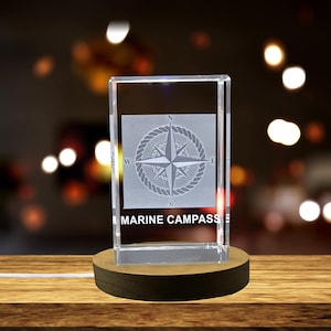 Op de afbeelding: Een heldere glazen kubus met een gegraveerd kompasontwerp aan de voorkant. Het kompas heeft een ster met vier punten en de letters N, S, E en W. De tekst "MARINE COMPASS" is onder het kompas gegraveerd. De kubus staat op een houten voet.