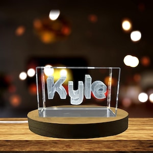 Peut inclure: Un bloc acrylique transparent avec le nom "Kyle" gravé, éclairé par une lumière chaude. Le bloc repose sur une base ronde en bois foncé. L'arrière-plan est flou avec des lumières bokeh.