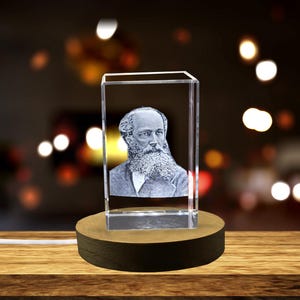 May include: A 3D crystal cube with a black and white engraved image of a man with a beard and mustache. The cube is on a wooden base.