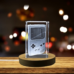 May include: A clear crystal cube with a 3D engraved image of a vintage Nintendo Game Boy handheld console. The cube is on a wooden base.