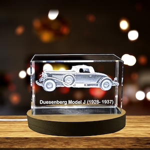 May include: A clear crystal cube with a 3D laser engraved image of a Duesenberg Model J car. The cube is on a wooden base. The text "Duesenberg Model J (1928-1937)" is engraved on the base of the cube.