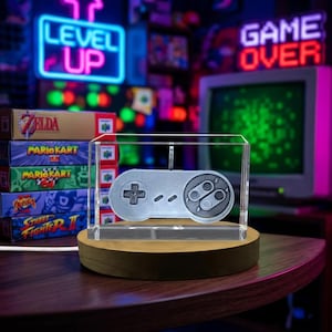 May include: A silver-toned video game controller ornament encased in a clear rectangular acrylic block, mounted on a circular wooden base. The controller features a directional pad on the left and two circular buttons on the right side.