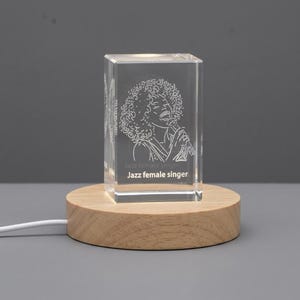 Könnte beinhalten: Ein klarer, rechteckiger Glasblock mit einer internen Lasergravur einer Sängerin mit lockigem Haar, die ein Mikrofon hält. Der Text "Jazz female singer" ist unten eingraviert. Der Block steht auf einem runden hellen Holzsockel.