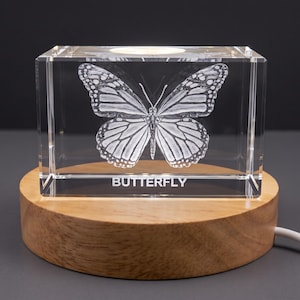 Könnte beinhalten: Ein klarer, rechteckiger Glasblock mit einem lasergravierten Monarchfalter und dem in Weiß gedruckten Wort BUTTERFLY. Der Block steht auf einem runden, hellen Holzsockel mit einem weißen Stromkabel an der Seite.