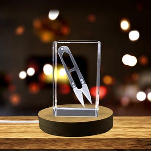 May include: A small, silver pair of thread snips are displayed in a clear glass cube on a dark wood base. The cube is illuminated from below, casting a soft glow on the snips.