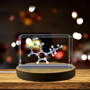 Puede incluir: Un modelo molecular encerrado en un bloque de acrílico transparente, exhibido sobre una base de madera. El modelo presenta una estructura compleja de esferas y varillas interconectadas, con un fondo borroso y cálido.