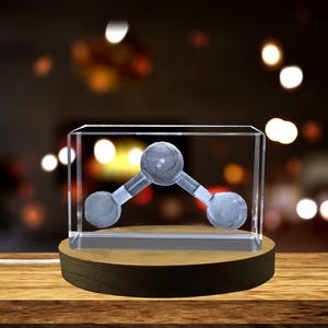 May include: A clear glass cube with a 3D etched image of a water molecule on a wooden base. The base is round and has a dark brown finish.
