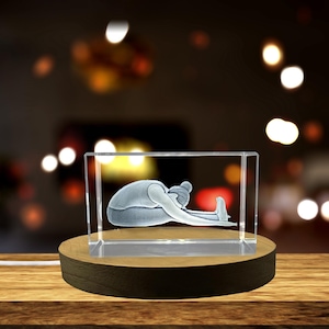 May include: A clear, rectangular glass block with a 3D etched image of a person in a seated forward bend yoga pose. The block sits on a round, wooden base with a warm light. The background is blurred with bokeh lights.