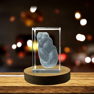 May include: A clear glass sculpture of a braided loaf of bread, encased in a rectangular block. The sculpture is displayed on a round wooden base with a dark brown edge. The background is blurred with warm, out-of-focus lights.