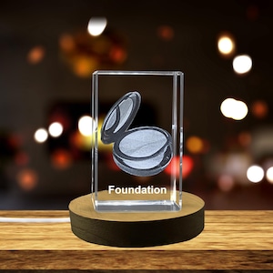 Può includere: Un cubo di vetro trasparente con un'immagine incisa in 3D di una cipria compatta. Il cubo è illuminato dal basso e poggia su una base di legno. Il testo "Foundation" è inciso sul fondo del cubo.