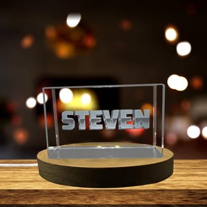Op de afbeelding: Een gepersonaliseerd LED-nachtlampje met de naam "STEVEN" verlicht in zilveren letters. Het licht is ingekapseld in een helder acrylblok, geplaatst op een houten voet. De achtergrond is wazig met bokeh-lichten.