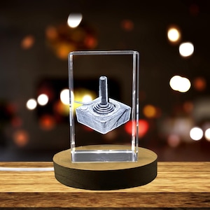 May include: A clear glass cube with a 3D etched image of a vintage video game joystick. The cube is on a wooden base with a white cord.