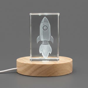 May include: A clear rectangular glass block featuring a laser-etched three-dimensional rocket ship with a plume of exhaust. The glass block is mounted on a round, light-colored wooden base with a white power cord extending from the side.