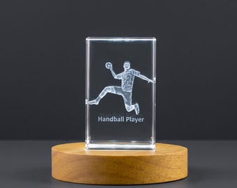 Può includere: Un blocco di vetro rettangolare trasparente con un'immagine 3D incisa al laser di un giocatore di pallamano in movimento aereo, con il testo "Handball Player" stampato sotto la figura, montato su una base circolare in legno chiaro.