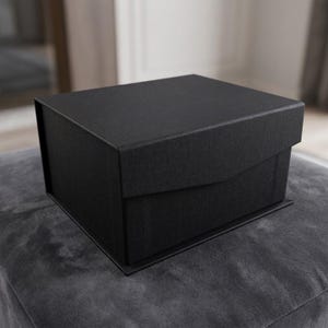 May include: A black, square gift box with a textured surface. The box has a lid that partially overlaps the base. The box is sitting on a gray, textured surface.