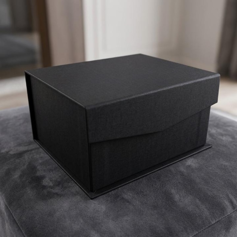 May include: A black, square gift box with a textured surface. The box has a lid that partially overlaps the base. The box is sitting on a gray, textured surface.