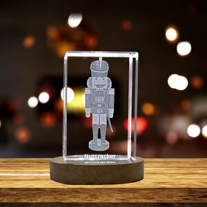 May include: A crystal figurine of a nutcracker soldier with a white base. The figurine is illuminated from within, casting a warm glow. The base of the figurine has the word "Nutcracker" inscribed on it.
