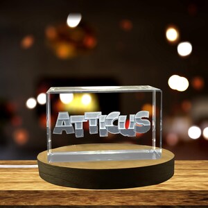 Könnte beinhalten: Ein klarer, rechteckiger Block mit dem Namen "Atticus" in silbernen und roten Buchstaben. Der Block steht auf einem runden Holzsockel. Der Hintergrund ist verschwommen mit warmen Lichtern.