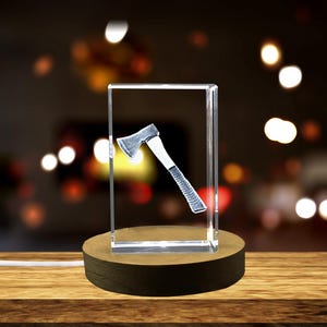 May include: A clear, rectangular glass block with a 3D axe design inside. The axe has a silver head and a white handle. The block sits on a round, dark brown wooden base.