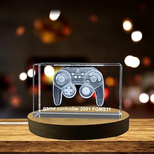 May include: A clear crystal cube with a 3D etched image of a video game controller. The cube is on a wooden base and the text "game controller 2001 FGMS11" is printed on the base.
