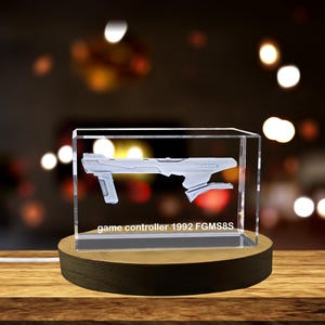 Op de afbeelding: Een transparante glazen kubus met een wit 3D-model van een gamecontroller erin. De kubus staat op een houten basis. De tekst "game controller 1992 FGMS8S" is op de basis gedrukt.
