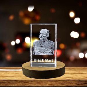 May include: A crystal cube with a 3D engraved image of a man with a mustache and beard. The cube is on a wooden base and the text "Black Bart" is engraved on the bottom of the cube.