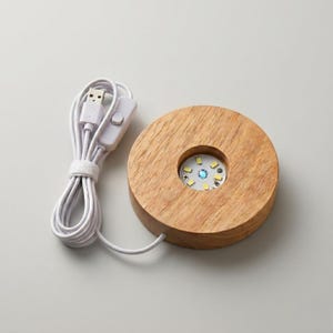 May include: A round, wooden LED light base with a USB cable. The base has a circular recess with a small LED light array. The light array emits a soft, multi-colored glow. The USB cable is white.