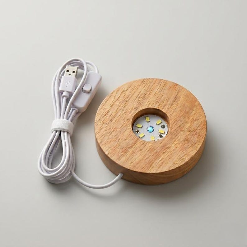 May include: A round, wooden LED light base with a USB cable. The base has a circular recess with a small LED light array. The light array emits a soft, multi-colored glow. The USB cable is white.