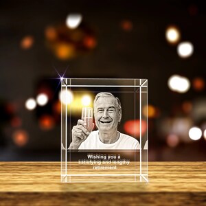 K&ouml;nnte beinhalten: Ein Kristallw&uuml;rfel mit einem Foto eines Mannes, der ein Glas Champagner h&auml;lt. Der Text "Wishing you a satisfying and lengthy retirement" ist auf der Unterseite des W&uuml;rfels graviert.