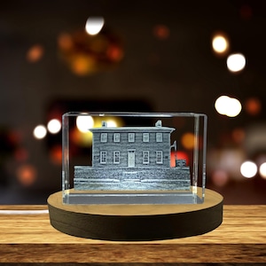 May include: A glass block featuring a detailed grey depiction of a building, set on a wooden base. The building is encased within the glass, and the base is illuminated. The background is blurred with bokeh lights.