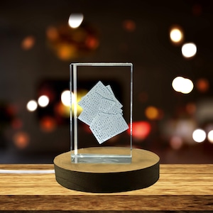 May include: A clear, rectangular glass block with a stack of textured, square-shaped objects inside, mounted on a round wooden base. The base is dark brown with a light brown top. The background is blurred with bokeh lights.