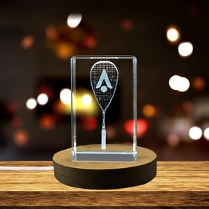 Könnte beinhalten: Ein Kristall-Briefbeschwerer mit einem 3D-geätzten Design eines Squashschlägers und eines Balls. Der Briefbeschwerer steht auf einem Holzsockel und wird von unten beleuchtet.