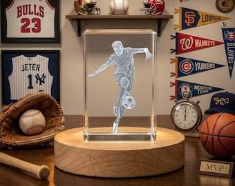 Può includere: Una scultura in vetro trasparente di un calciatore in azione, montata su una base di legno. Lo sfondo include cimeli sportivi come palloni da baseball, caschi e maglie incorniciate. Sono visibili anche un pallone da basket e un cronometro.