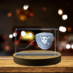 Op de afbeelding: Een heldere glazen kubus met een houten basis, waarop een wit gegraveerd piratenooglapje met een schedel en gekruiste beenderen is afgebeeld.