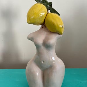 Puede incluir: Una escultura de cerámica de un torso femenino con dos limones amarillos y hojas verdes en lugar de una cabeza. La escultura es de color rosa claro y se encuentra sobre una superficie turquesa.