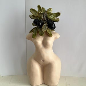 Puede incluir: Escultura de cerámica de un torso femenino con una cabeza de hojas verdes y aceitunas negras. La escultura es de color beige claro y tiene un acabado liso. El tocado de rama de olivo añade un elemento natural a la obra.