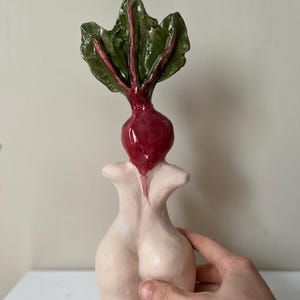 Puede incluir: Una escultura de cerámica de una remolacha con hojas verdes y una raíz roja. La escultura está unida a una forma estilizada de color nude. La escultura está sostenida por una mano.
