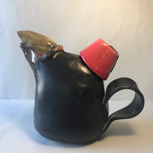 Puede incluir: Una tetera de cerámica negra con una tapa roja en forma de fez. La tetera tiene una forma única y orgánica y un asa que se extiende desde el lateral.