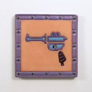 May include: A purple ceramic tile with a light orange background and a blue and black stylized image of a toy gun.