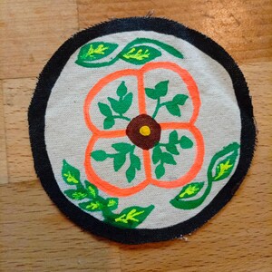 Puede incluir: Parche de tela redondo con diseño floral. El parche presenta un fondo color crema con un contorno de flor naranja, hojas verdes y un centro marrón con un punto amarillo. Está bordeado por un borde cosido negro.