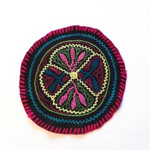 Op de afbeelding: Een ronde, zwarte stoffen onderzetter met een kleurrijk geborduurd ontwerp. Het ontwerp heeft een centrale bloem met vier bloemblaadjes, elk met een andere kleur en patroon. De onderzetter is omgeven door een roze, blauwe en groene rand.