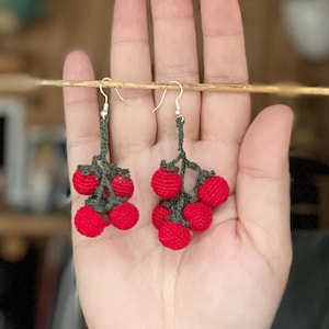 Op de afbeelding: Een paar handgemaakte gehaakte oorbellen met clusters van rode bollen, die op bessen lijken, bevestigd aan groene stelen. De oorbellen hangen aan zilverkleurige haken. De achtergrond is wazig, wat op een natuurlijke omgeving duidt.