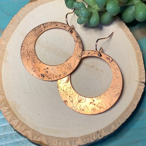 Puede incluir: Un par de pendientes de aro de cobre con un diseño floral. Los pendientes están sobre una superficie de madera.