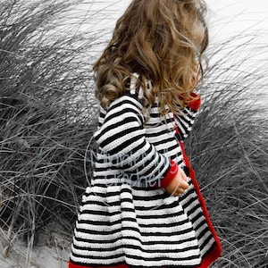 May include: A young girl wearing a black and white striped cardigan with red trim. The cardigan has a flared skirt and is crocheted. The girl is wearing red boots and is standing in a field of tall grass.